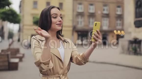 一个长着棕色头发戴着米色壕沟的漂亮女孩打着智能手机微笑挥手视频的预览图