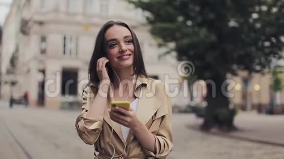 漂亮的女孩戴着耳机听音乐看着她的智能手机和微笑站在城市里视频的预览图