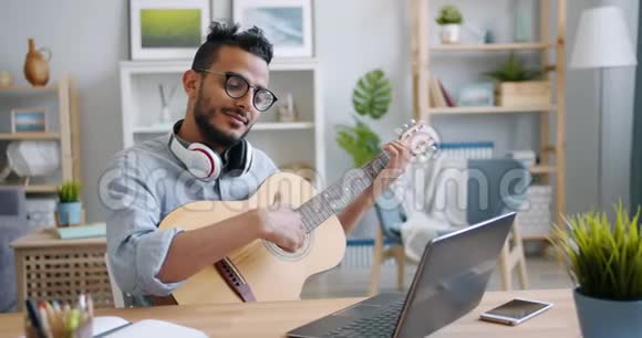 年轻的阿拉伯人弹吉他在公寓的桌子上使用笔记本电脑视频的预览图