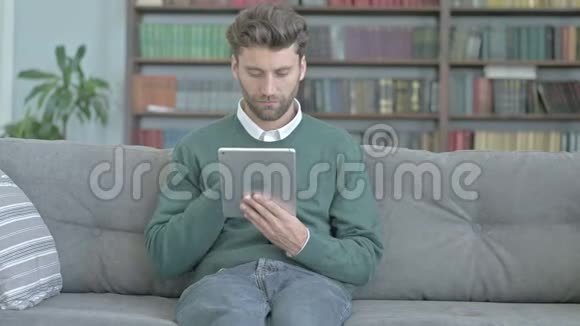 放松的男人坐在沙发上一边在平板电脑上滚动一边微笑视频的预览图