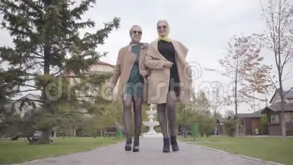 两位穿着秋色米色外套短裙和太阳镜的白种人成熟女性在城市公园散步视频的预览图