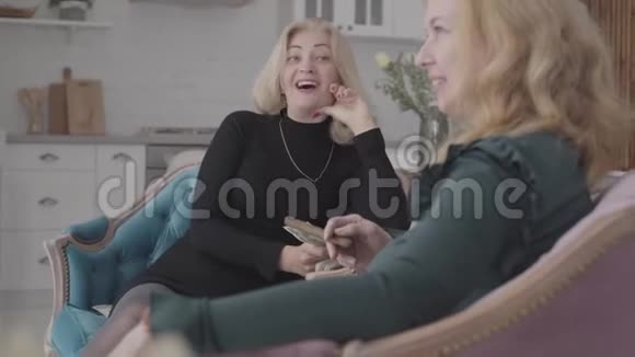 两个快乐成熟的高加索女人坐在舒适的扶手椅上拿着钱和雪茄聊天老年女性视频的预览图
