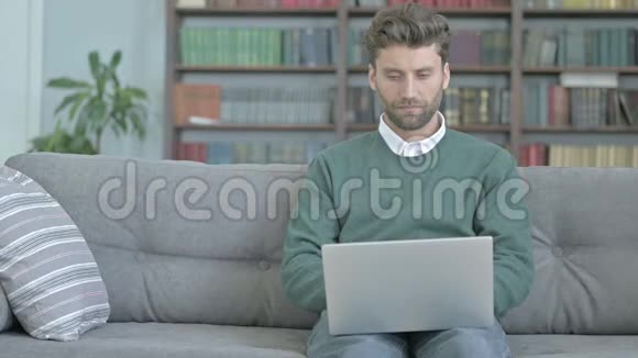兴奋的年轻人坐在沙发上一边用笔记本电脑一边庆祝视频的预览图
