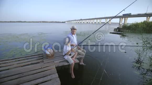 快乐的家庭父亲和他戴着草帽的小男孩在河边的码头上钓鱼玩得很开心视频的预览图