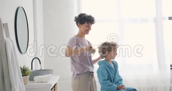 穿着睡衣梳着小男孩的头发的年轻女子微笑着视频的预览图
