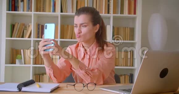 年轻有魅力的女商人坐在笔记本电脑前摆姿势在手机上自拍的特写视频的预览图
