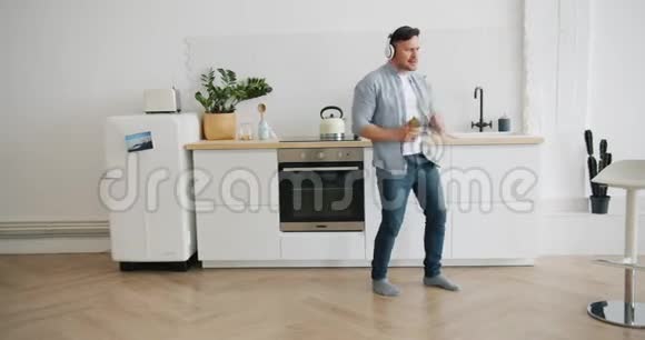 戴着耳机听音乐的男人在厨房里用智能手机跳舞视频的预览图