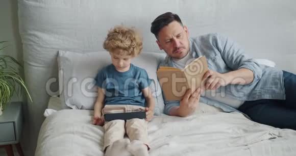 严肃的家伙在床上看书而小孩子在平板电脑上看动画片视频的预览图