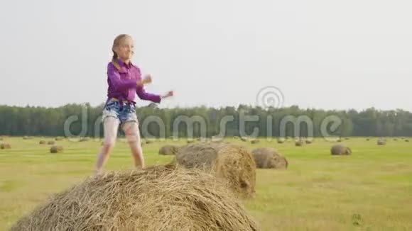 快乐的女孩在收获场的干草堆上跳舞在乡下的干草堆上跳舞的快乐女孩青年视频的预览图