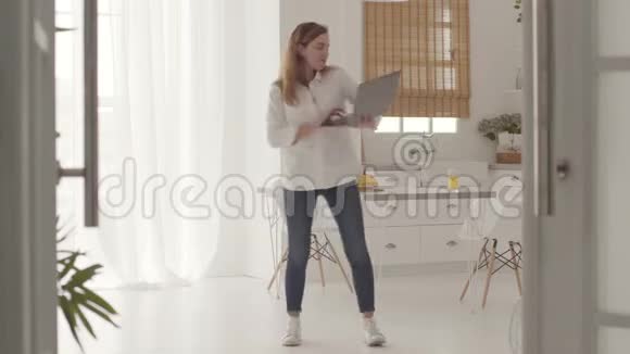 年轻的白人女孩穿着休闲服装在轻盈的现代厨房里和笔记本电脑跳舞快乐美丽的女人视频的预览图