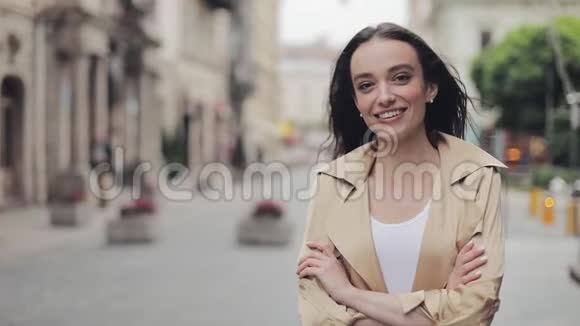 穿着米黄色壕沟的漂亮女孩的肖像微笑着笑着交叉着双手站在城市里视频的预览图