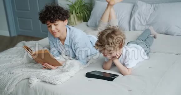 小男孩在平板电脑上看动画片而妈妈则在床上看书视频的预览图