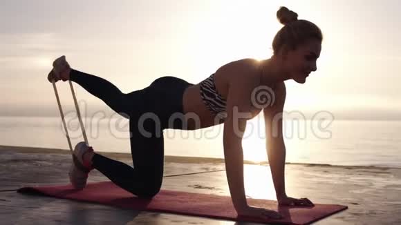 穿着运动服装的白种人运动服的女人在瑜伽地毯上用弹性带和伸展的腿以及靠近大海的全身视频的预览图