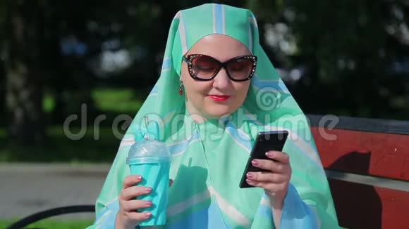 一位戴着浅色围巾和墨镜的年轻穆斯林妇女坐在公园的长凳上喝着咖啡在公园里聊天视频的预览图