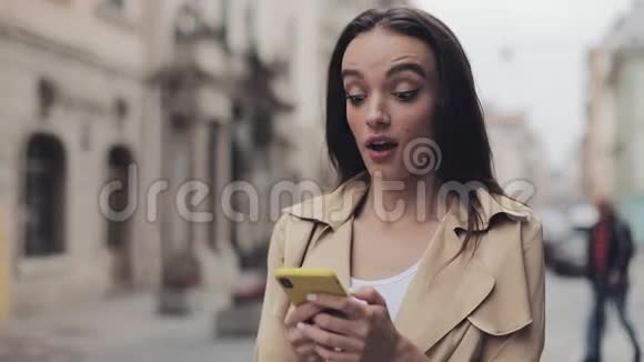 年轻迷人的女孩看起来非常印象深刻拿着手机聊天和笑说哇站在城市视频的预览图