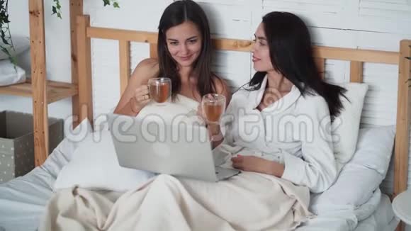 两个女朋友坐在床上喝茶在笔记本电脑上看有趣的电影笑视频的预览图