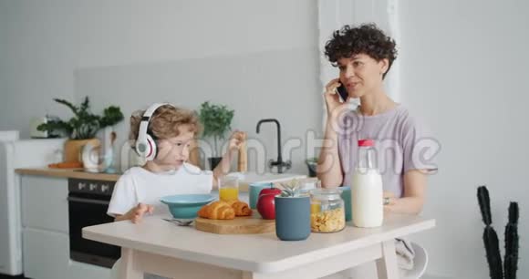 女人一边打手机一边儿子在厨房里用耳机欣赏音乐视频的预览图