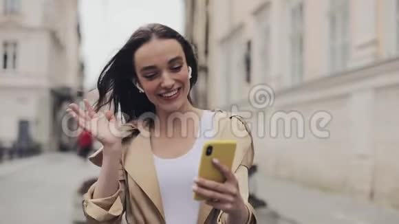 美丽的快乐女孩戴着无线耳机打着视频电话微笑着挥手散步视频的预览图
