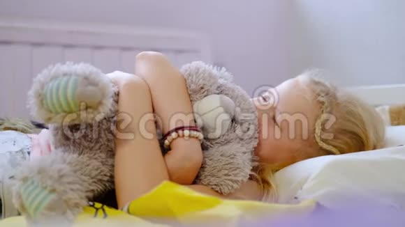 漂亮的金发小女孩在床上醒来一大早就对着镜头微笑女孩拥抱玩具熊视频的预览图