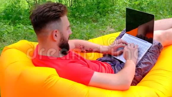 穿红色T恤的家伙躺在空中沙发上在笔记本电脑上工作有胡子的商人自由职业者或游戏玩家视频的预览图