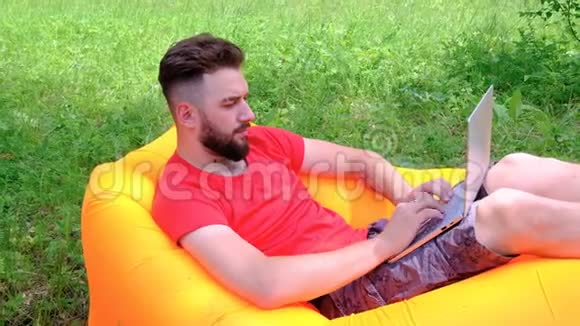 穿红色T恤的人躺在充气床垫上在笔记本电脑上工作有胡子的年轻人是个自由职业者视频的预览图