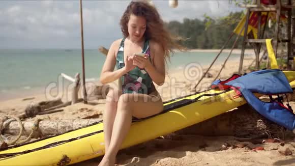 穿着泳装坐在沙滩上用智能手机微笑的女人视频的预览图