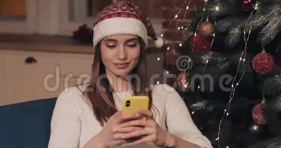 戴着圣诞帽坐在圣诞树旁的美丽的年轻白种人女士使用她的智能手机概念视频的预览图