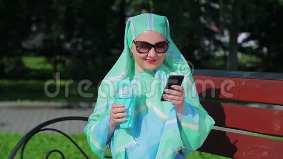 一位戴着浅色围巾和太阳镜的年轻穆斯林妇女坐在公园的长凳上喝着咖啡说话视频的预览图