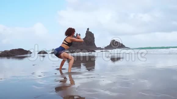 美丽的运动女孩穿着运动的衣服在海滩上表演蹲健康的生活方式健身视频的预览图