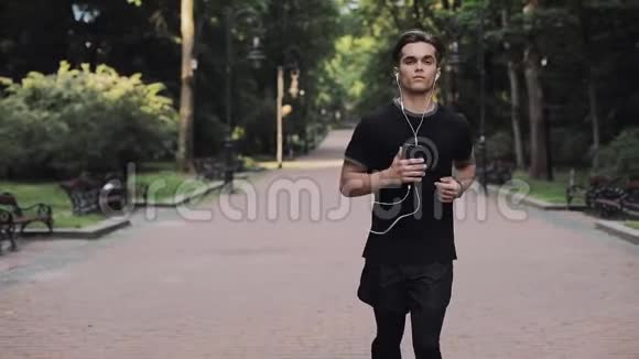 戴着耳机戴着智能手机手里拿着慢跑服在公园里跑步的帅哥视频的预览图