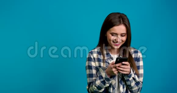 微笑女孩在格子衬衫的正面视图使用智能手机隔离在蓝色背景看镜头视频的预览图