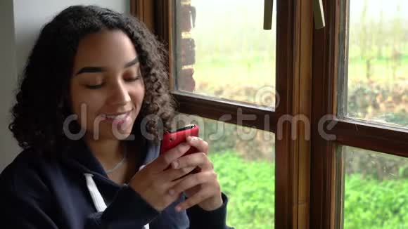 一位非洲裔美国少女使用手机或智能手机在社交媒体或Texti上坐在窗边视频的预览图