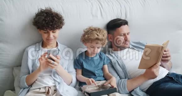 母亲用智能手机儿子拿着平板电脑父亲在家里的床上看书视频的预览图