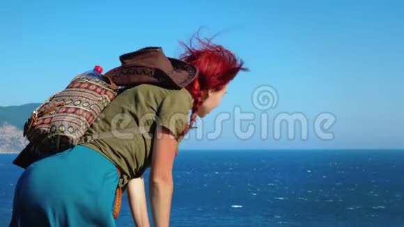 一个年轻的红发女孩旅行者戴着牛仔帽和背包站在山顶上俯视大海视频的预览图