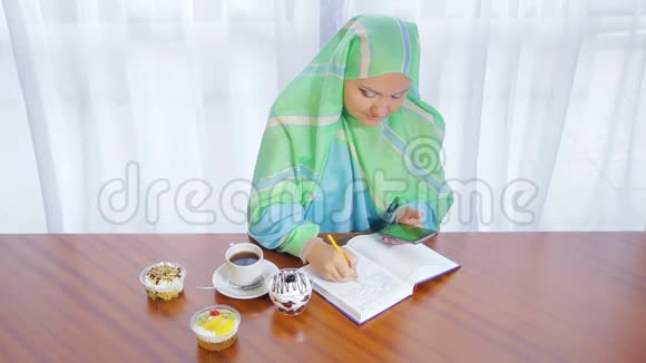 一个年轻的穆斯林妇女在一家咖啡馆里戴着一条浅色围巾用电话日记和喝茶远程工作视频的预览图