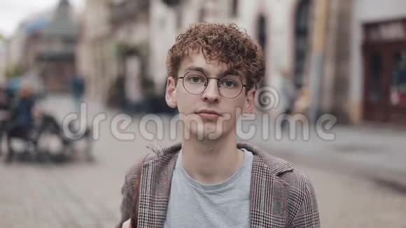 在旧城街道上一个戴着眼镜和夹克看着摄像机的书呆子青年的肖像视频的预览图