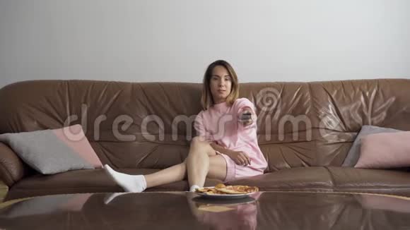无聊的年轻女人坐在棕色的皮革沙发上看电视使用远程的可爱女性切换通道视频的预览图