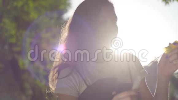 十几岁的女孩在阳光下在公园的相机上吹肥皂泡可爱的年轻女子独自在户外活动视频的预览图