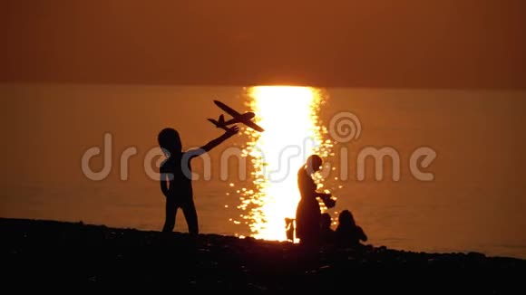 一个快乐的孩子在日落海边玩玩具飞机的剪影家庭幸福视频的预览图