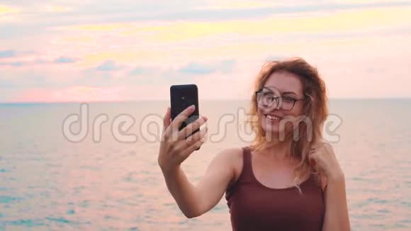 穿着棕色t恤的可爱年轻迷人的女士在冰冷的大海和粉红色的日落前拍摄她的脸女孩和视频的预览图