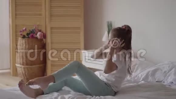 有趣可爱的小女孩带着耳机在床上听音乐年轻的音乐爱好者在家玩音乐视频的预览图