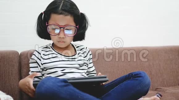 亚洲小女孩在沙发上用平板电脑放松视频的预览图