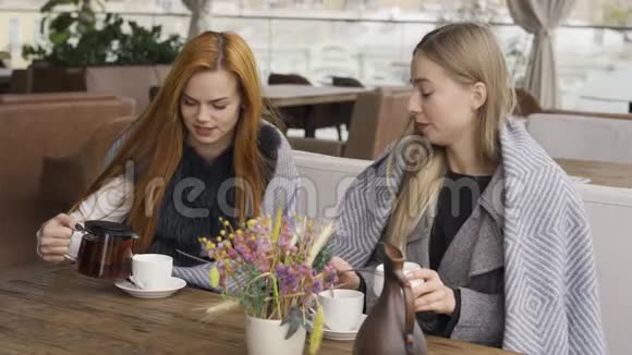 两个裹着毯子的白人女友坐在咖啡馆里聊天喝着热茶年轻女性放松视频的预览图