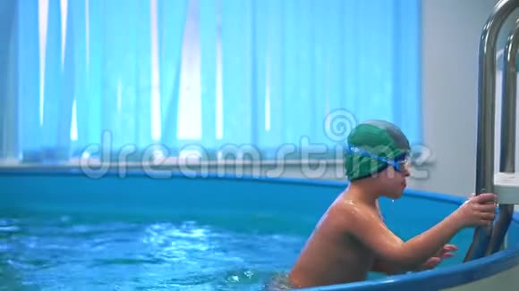 一个男孩在游泳池里带着泳镜上楼梯走出游泳池视频的预览图