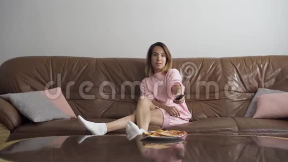 漂亮的年轻女人坐在家里的棕色皮革沙发上看电视使用远程的可爱女性切换通道视频的预览图