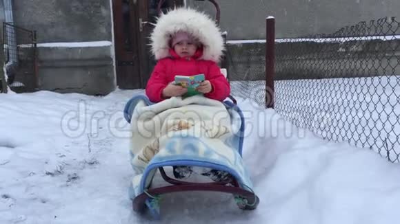 小女孩坐在雪橇上躲在毯子里抓住飞舞的雪花看书视频的预览图
