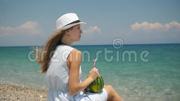 年轻美丽的欧洲女孩戴着白帽子在海滩上喝着新鲜的水果鸡尾酒视频的预览图