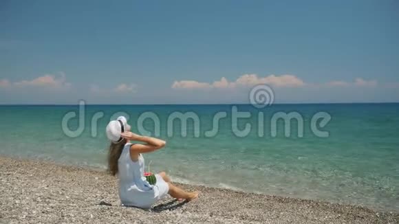 年轻美丽的白种人女孩戴着白帽子独自坐在海滩上海岸景色视频的预览图