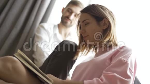 漂亮的年轻夫妇正坐在家的窗边一个女孩正在给她的男朋友或丈夫读一本书视频的预览图