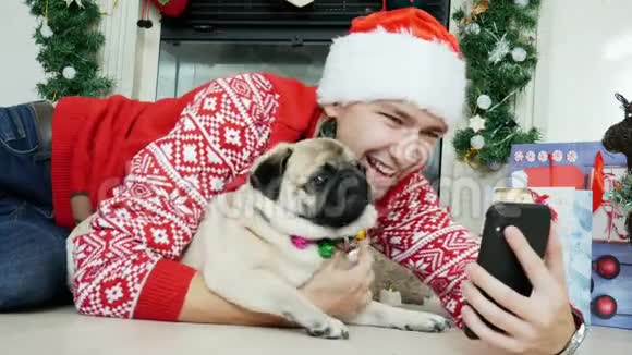 男人抱着滑稽的哈巴狗穿着圣诞服装戴着圣诞帽用手机新年和圣诞节视频的预览图